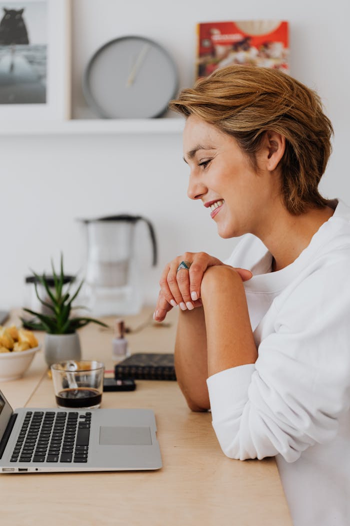 Woman in white top smiling while using a laptop at a bright home desk. Relaxed work atmosphere.